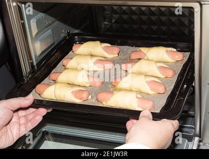 Femme insère une plaque de cuisson de saucisses dans la pâte dans le four. Cuisson au four. Banque D'Images