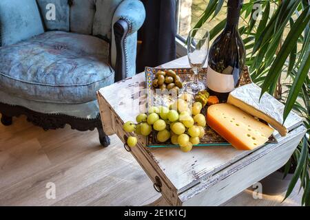 plateau de fromages avec raisins et vin sur une table près d'une fenêtre avec une chaise confortable. Décoration intérieure moderne Banque D'Images