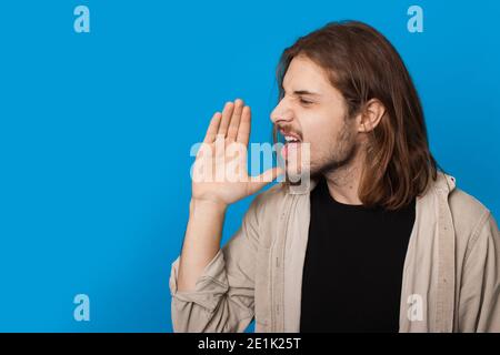 L'homme caucasien avec la barbe et les cheveux longs crie dessus un mur de studio bleu avec espace libre annonçant quelque chose Banque D'Images