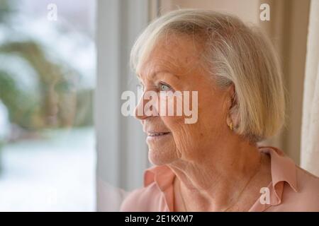 Portrait d'une femme âgée attrayante debout à l'intérieur à la maison regarder par une fenêtre avec une expression réfléchie Banque D'Images