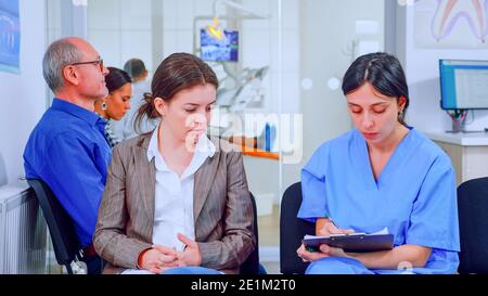 Une infirmière prend des notes sur le presse-papiers au sujet des problèmes dentaires des patients qui attendent un orthodontiste assis sur une chaise dans la salle d'attente de la clinique stomatologique. Assistant expliquant la procédure médicale à la femme. Banque D'Images