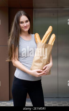 Jeune femme caucasienne en vêtements décontractés portant un sac en papier avec baguette, se tenir souriant devant l'ascenseur de condominium. Nourriture et colis deli Banque D'Images