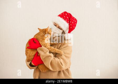 fille dans un chapeau de père noël et un masque médical de protection tient un chat gras de gingembre dans ses mains. Nouvelle année 2021 Banque D'Images