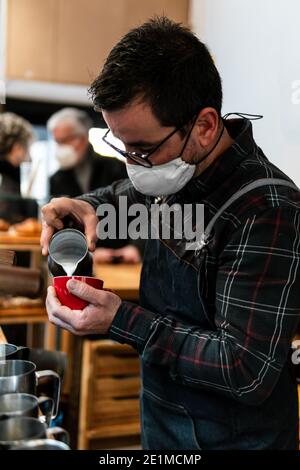 Barista caucasien avec masque versant du lait dans une tasse de café rouge dans un café, devant la machine à café avec deux clients non focalisés à l'arrière Banque D'Images