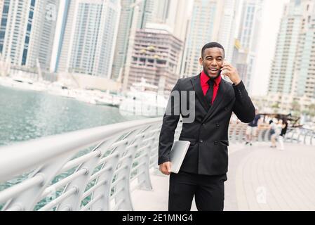 Jeune homme d'affaires afro-américain debout à Dubai Marine et parlant sur son téléphone portable avec le sourire sur son visage. Banque D'Images