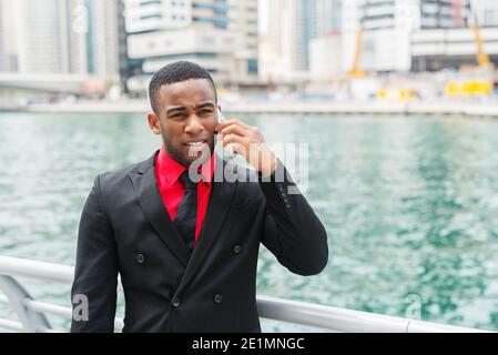 Jeune homme d'affaires afro-américain debout à Dubai Marine et parlant sur son téléphone portable avec un regard inquiet sur son visage. Banque D'Images