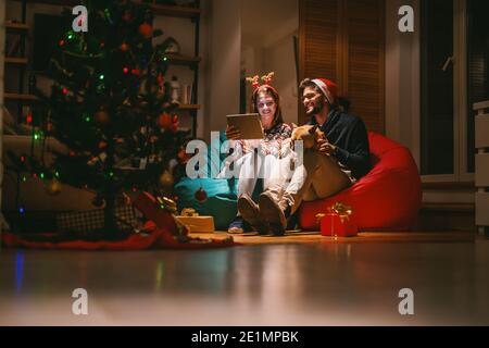 Couple utilisant une tablette tout en étant assis dans le salon. Homme tenant le chien. Sur les têtes chapeau de père noël et serre-tête de renne. Arbre de Noël en premier plan. Noël Banque D'Images