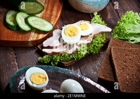 préparation d'un sandwich à la viande avec des feuilles de laitue verte, des œufs hachés, des concombres et du pain de seigle ou de blé, sur fond de table rustique foncé en bois, personne Banque D'Images