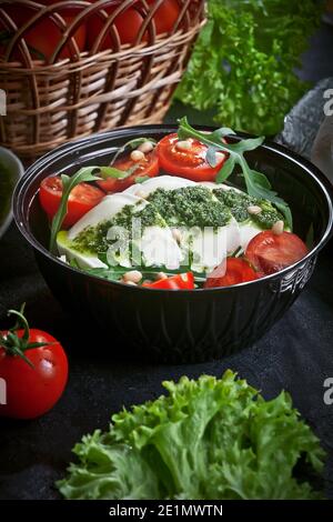 salade de mozzarella fraîche avec laitue et feuilles d'arugula, sauce pesto, tomates cerises fraîches et noix de cèdre, sur une assiette noire sur table d'ardoise sombre Banque D'Images