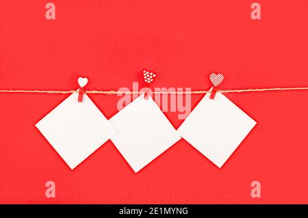 trois autocollants carrés blancs vides pour les notes épinglées à la corde avec une épingle à linge avec des coeurs sur fond rouge, concept de jour de saint valentin, carte de voeux Banque D'Images