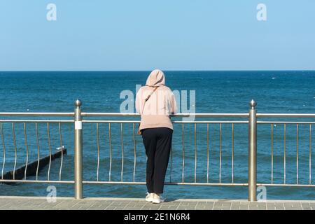 Femme en survêtement se tient avec son dos sur la mer et regarde dans la distance. Concept d'attente, adieu à la mer. Mise au point sélective. Banque D'Images