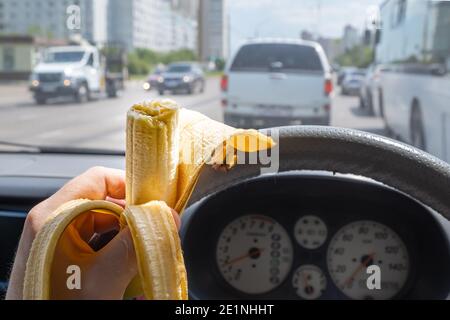 Nourriture, banane dans la main d'un conducteur conduisant une voiture qui conduit sur une rue de ville parmi la circulation de la ville Banque D'Images