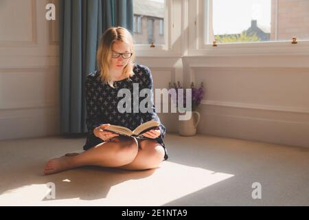 Une jeune femme lit un livre dans la lumière naturelle de la baie vitrée de son appartement d'Édimbourg. Banque D'Images