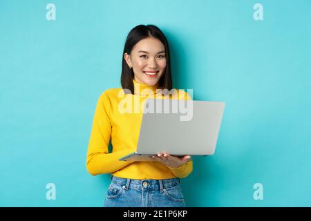 Femme asiatique heureuse en pull jaune utilisant un ordinateur portable, faire des achats en ligne ou travailler, debout sur fond bleu Banque D'Images