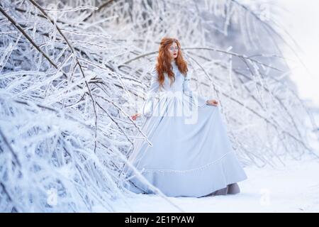 Une jeune femme à tête rouge, une princesse, marche dans une forêt d'hiver en robe bleue. Banque D'Images