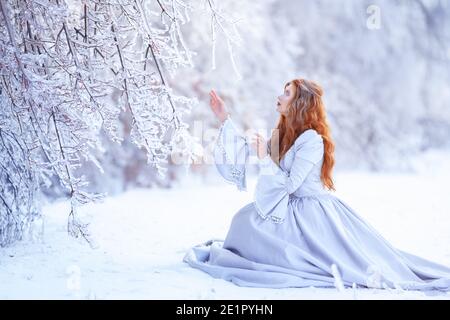 Une jeune femme à tête rouge, une princesse, marche dans une forêt d'hiver en robe bleue. Banque D'Images