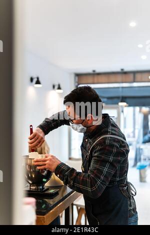 Boulanger caucasien avec masque préparant un gâteau dans une boulangerie. Il mélange les ingrédients dans un bol. Banque D'Images