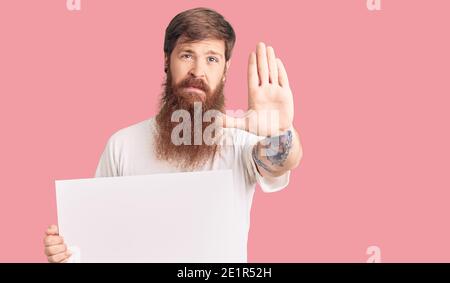 Beau jeune homme à tête rouge avec une longue barbe tenant une bannière vide vierge avec main ouverte faisant signe d'arrêt avec une expression sérieuse et confiante, défense g Banque D'Images