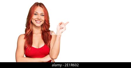 Jeune femme à tête rouge portant des vêtements décontractés avec un grand sourire sur le visage, pointant avec la main et le doigt vers le côté regardant l'appareil photo. Banque D'Images