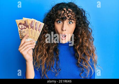 Jeune fille hispanique détenant 5000 billets de forint hongrois effrayé et stupéfié par la bouche ouverte pour la surprise, le visage incrédulé Banque D'Images