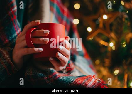 Femme méconnue tenant une tasse rouge confortable sur fond d'arbre de Noël brillant. Fille avec boisson chaude - thé, café ou cacao. Concept de la nouvelle année Banque D'Images