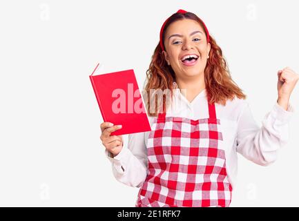 Jeune femme latine portant un tablier tenant livre de recettes criant fier, célébrant la victoire et le succès très excité avec les bras levés Banque D'Images