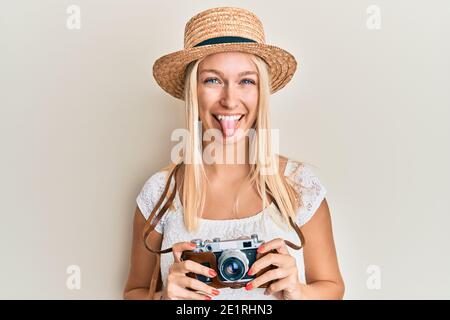 Jeune fille blonde portant un chapeau d'été en utilisant la caméra collant la langue dehors heureux avec l'expression drôle. Banque D'Images