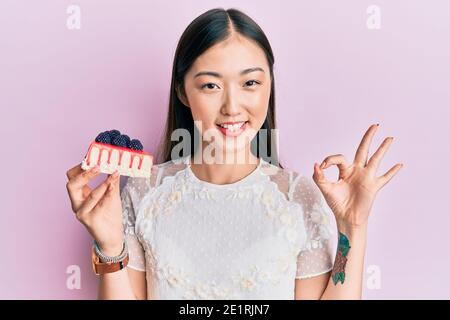 Jeune femme chinoise mangeant cheesecake faisant signe ok avec les doigts, souriant sympathique gesturant excellent symbole Banque D'Images