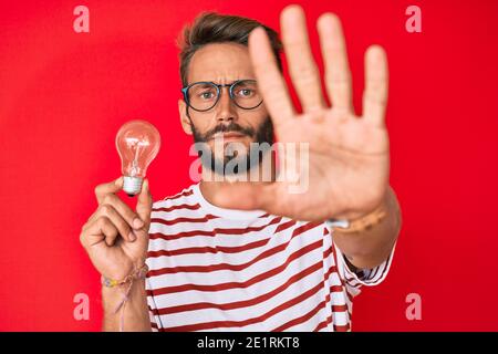 Beau caucasien homme avec barbe tenant ampoule pour l'inspiration et l'idée avec main ouverte faisant stop signe avec une expression sérieuse et confiante, def Banque D'Images