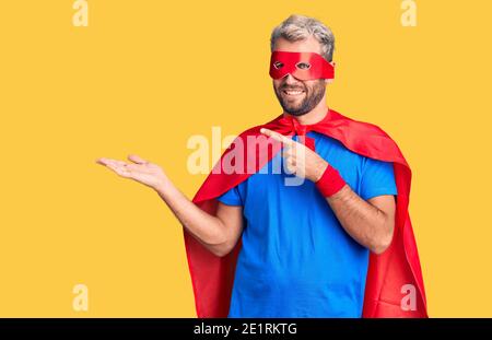 Jeune homme blond portant un super-héros crème émerveillé et souriant à l'appareil photo tout en présentant avec la main et en pointant avec le doigt. Banque D'Images