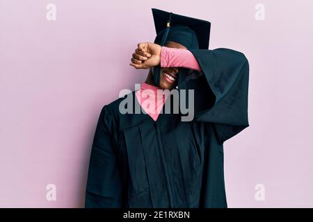 Jeune fille afro-américaine portant un chapeau de remise de diplôme et une robe de cérémonie couvrant les yeux avec le bras souriant gai et drôle. Concept aveugle. Banque D'Images