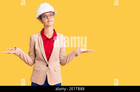 Jeune belle femme portant un architecte hardhat expression sans indice et confuse avec les bras et les mains soulevées. Doute concept. Banque D'Images