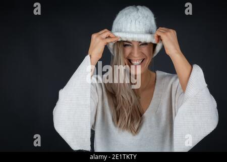 Femme blonde joueur tirant vers le bas tricoté chapeau blanc et sourire, fond sombre Banque D'Images