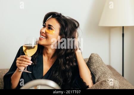 Vue latérale d'une heureuse femme brune se relaxant à la maison sur un canapé. Femme souriante dans un peignoir en satin tenant le vin. Banque D'Images