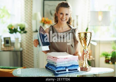 Portrait d'une femme de ménage souriante et tendance de 40 ans avec blouse en soie et pantalon beige avec fer à vapeur, planche à repasser, pile de vêtements pliés repassés et ged Banque D'Images