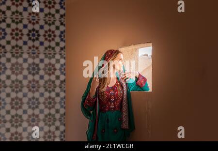 magnifique fille timide dans les vêtements traditionnels omanais regardant à travers le fenêtre et couvrant son visage avec un voile pour se cacher les yeux des gens qui marchent à proximité Banque D'Images