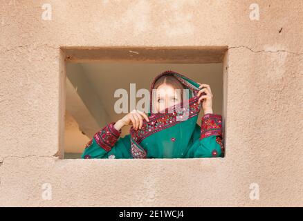 magnifique fille timide dans les vêtements traditionnels omanais regardant à travers le fenêtre et couvrant son visage avec un voile pour se cacher les yeux des gens qui marchent à proximité Banque D'Images