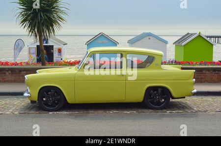 Voiture classique jaune Ford Anglia sur la route du front de mer en face des cabines de plage. Banque D'Images