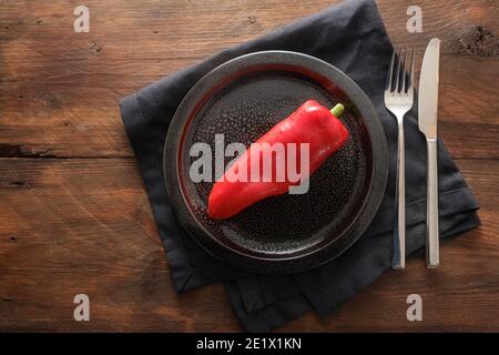 Poivron rouge brut pointu sur une assiette sombre, couverts et serviette sur un fond rustique en bois, un concept alimentaire sain ou un régime de légumes à perdre de poids Banque D'Images