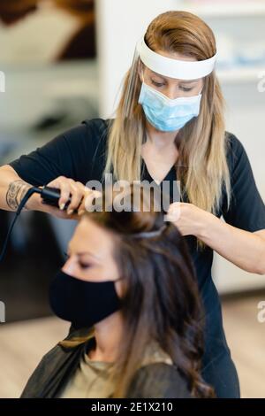 Coiffeur, protégé par un masque, peignant les cheveux de son client avec un fer à repasser dans un salon. Banque D'Images