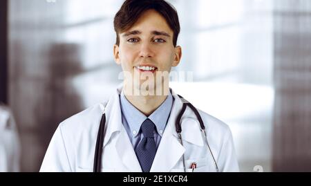 Jeune homme sympathique médecin debout avec presse-papiers dans la clinique moderne. Une collègue de sexe féminin est occupée à remplir le formulaire de dossiers de traitement sur le Banque D'Images