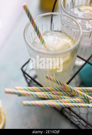 Vue rapprochée d'une tranche de citron et de petits cubes de glace autour dans un verre de limonade. D'autres pailles colorées sont au fond. Banque D'Images