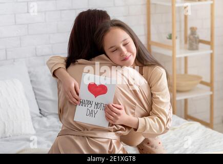 Adorable jeune fille embrassant sa mère, la félicitant de la fête des mères, tenant une carte de voeux sur le lit à la maison Banque D'Images