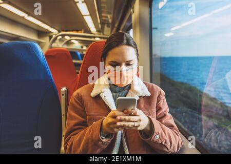 Une fille dans un masque facial de protection sur le train utilise un téléphone cellulaire. Voyage en train ou en train de banlieue. Banque D'Images