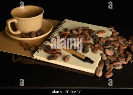 De nombreux grains de cacao aromatiques entiers, ovales et bruns, ainsi qu'une tasse à thé et une soucoupe sont situés sur une feuille vierge d'un carnet ouvert, et un stylo à bille se trouve Banque D'Images