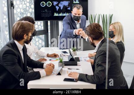 Homme mature et confiant réalisateur dans un masque de protection du visage, salutation amicale avec le poing frapper chaque membre de son équipe multiethnique, pendant les affaires Banque D'Images