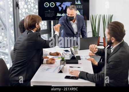 Homme mature et confiant réalisateur dans un masque de protection du visage, salutation amicale avec le poing frapper chaque membre de son équipe multiethnique, pendant les affaires Banque D'Images