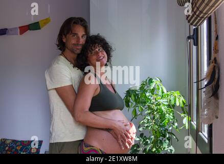 Homme à cheveux longs tenant le ventre de la femme enceinte heureuse par la fenêtre de la chambre. S'attendre aux parents, à la nouvelle vie, à la famille, aux concepts de la fête des mères Banque D'Images
