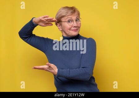 Femme caucasienne senior gestante avec les mains montrant le grand et grand signe, symbole de mesure souriant regardant la caméra. Studio tourné sur un mur jaune. Banque D'Images