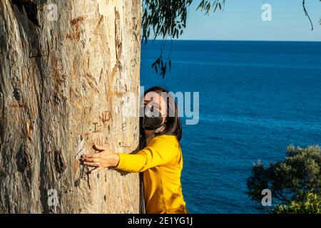 Femme avec masque noir qui embrasse le tronc d'un grand eucalyptus avec la mer en arrière-plan Banque D'Images